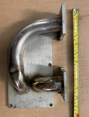 Original welded supercharger manifold on a metal plate, with a tape measure alongside for scale